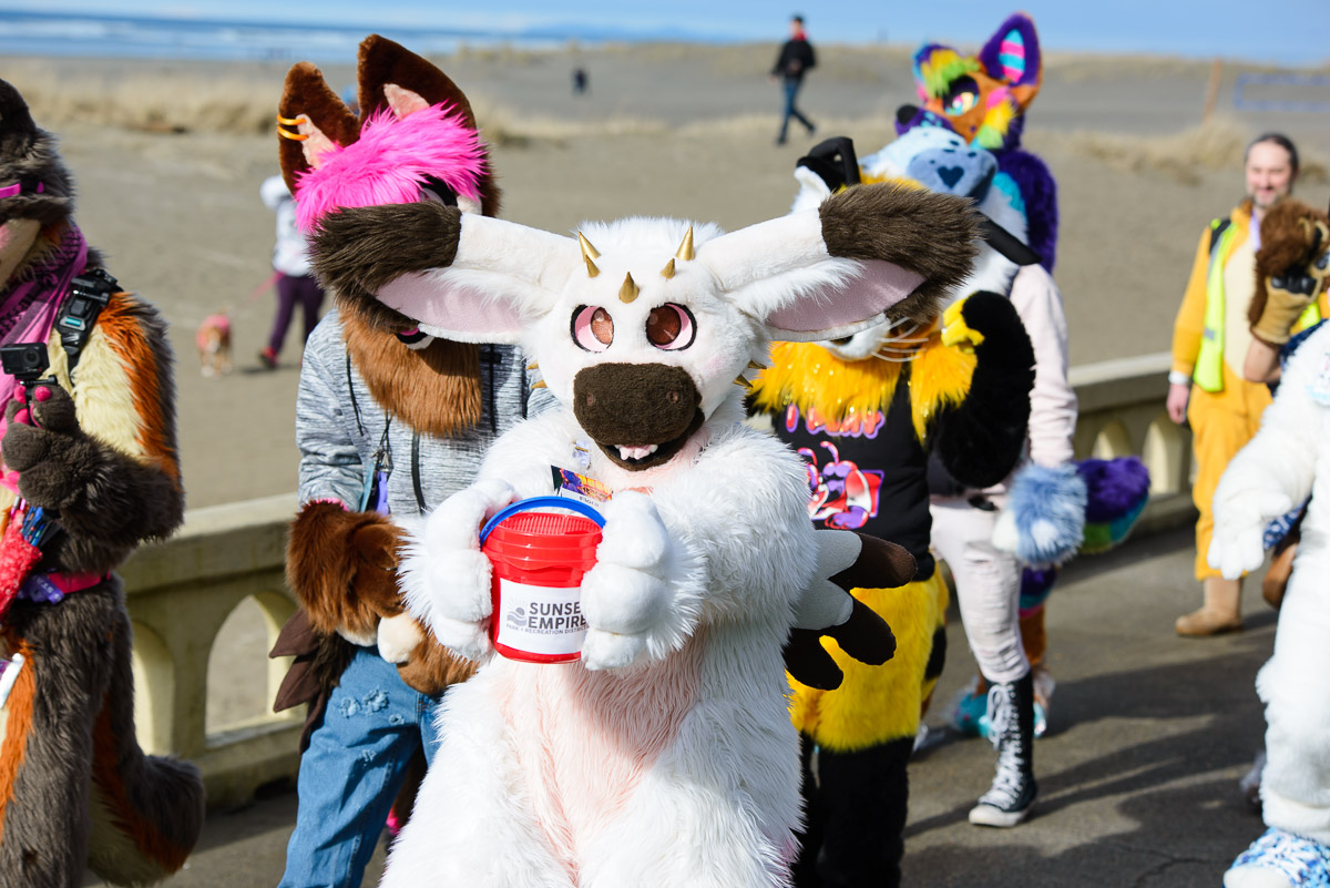 A picture of a white dutch angel dragon fursuiter holding out one of the donation buckets in the 2025 Fursuit Walk