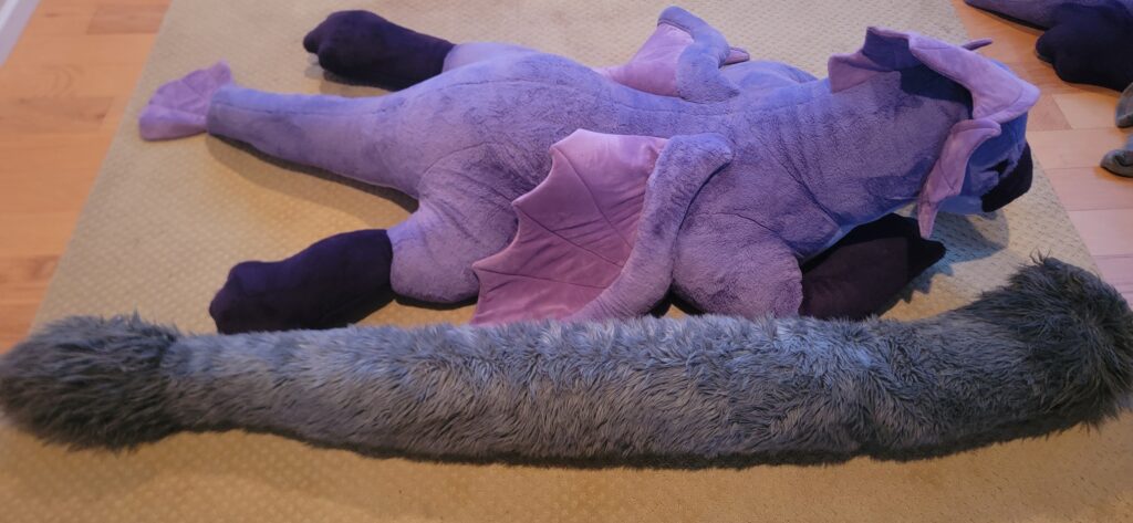 A picture of the grey fursuit tail next to the giant Finneus plush to give a sense of scale. The tail is just as long as Finneus