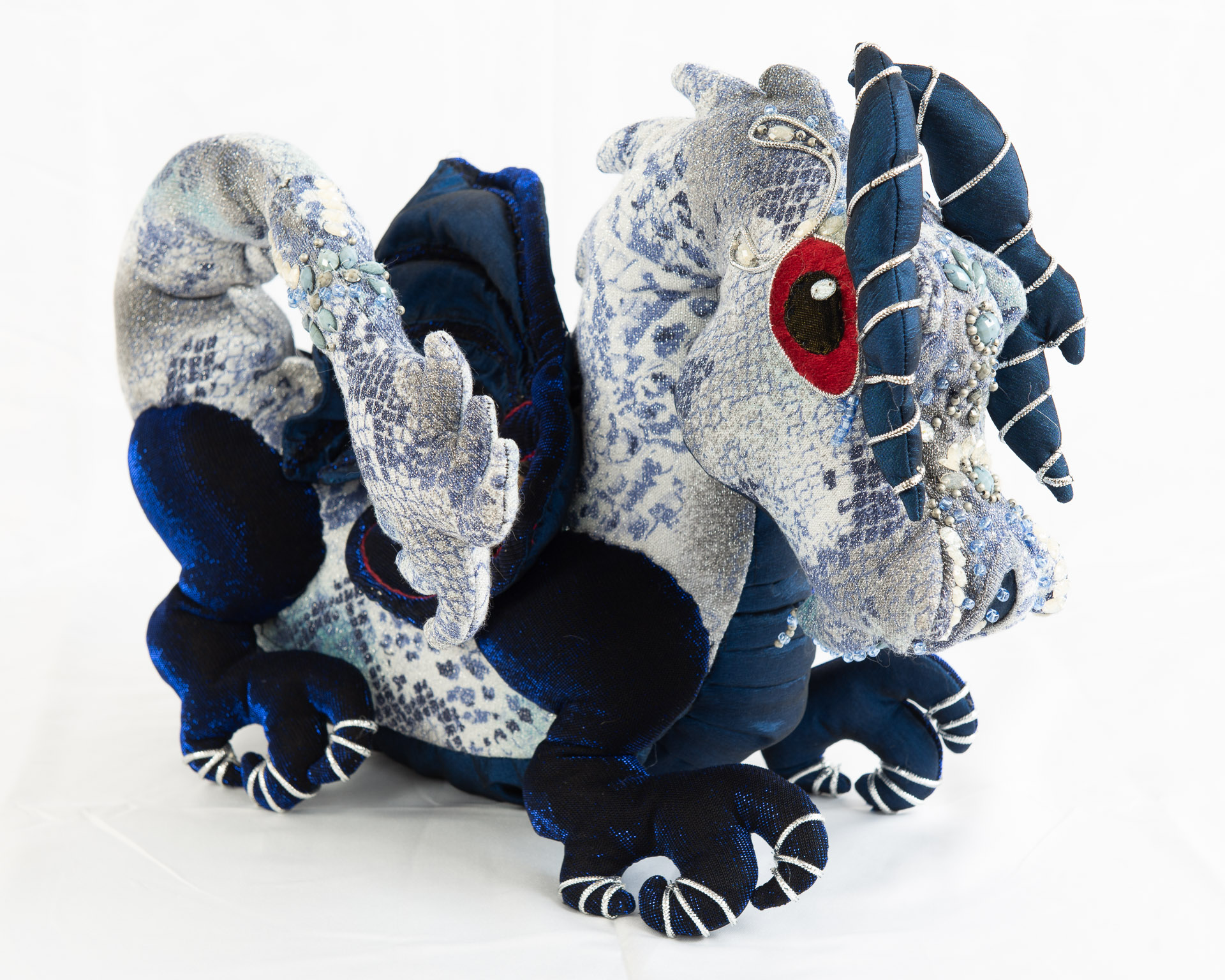 An intricate handcrafted dragon plush, it has striking red eyes, a silver body with different fabrics used to create scale effects, and deep blue limbs. 