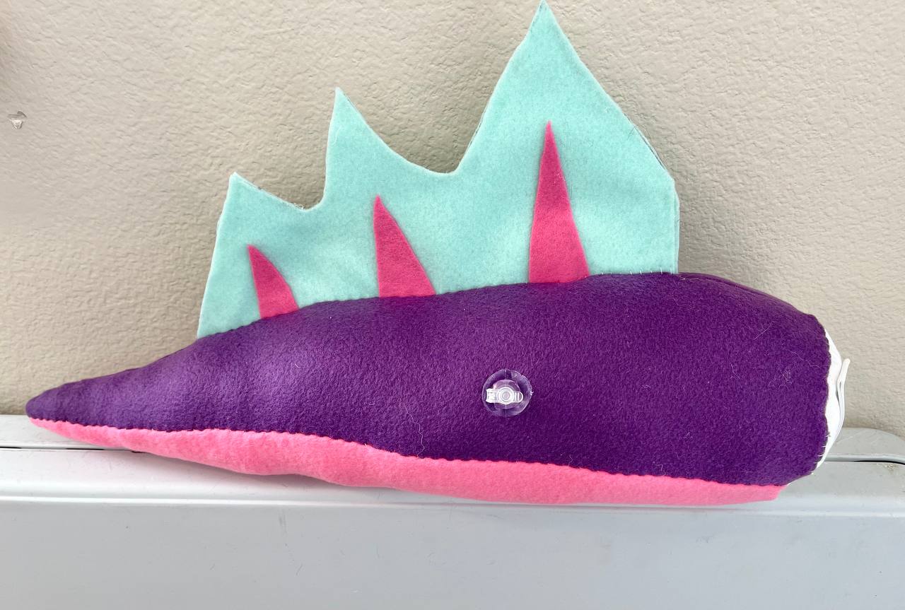 Picture of a Pooltoy inspired fursuit tail, made by Radio Neyo. It is mainly purple with a pink underside and light blue and pink fins on the top. 