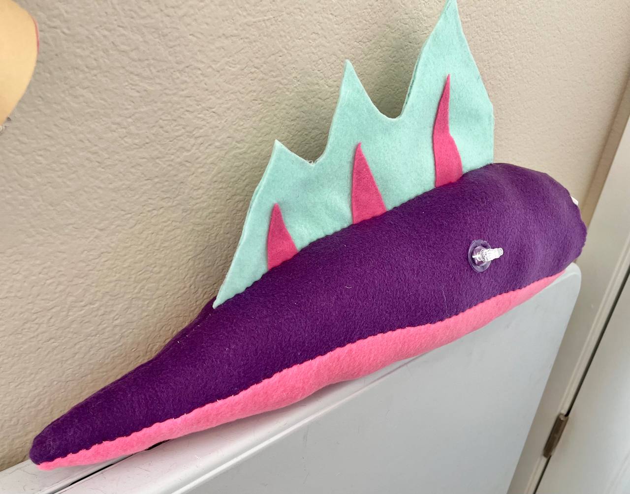 Picture of a Pooltoy inspired fursuit tail, made by Radio Neyo. It is mainly purple with a pink underside and light blue and pink fins on the top. 