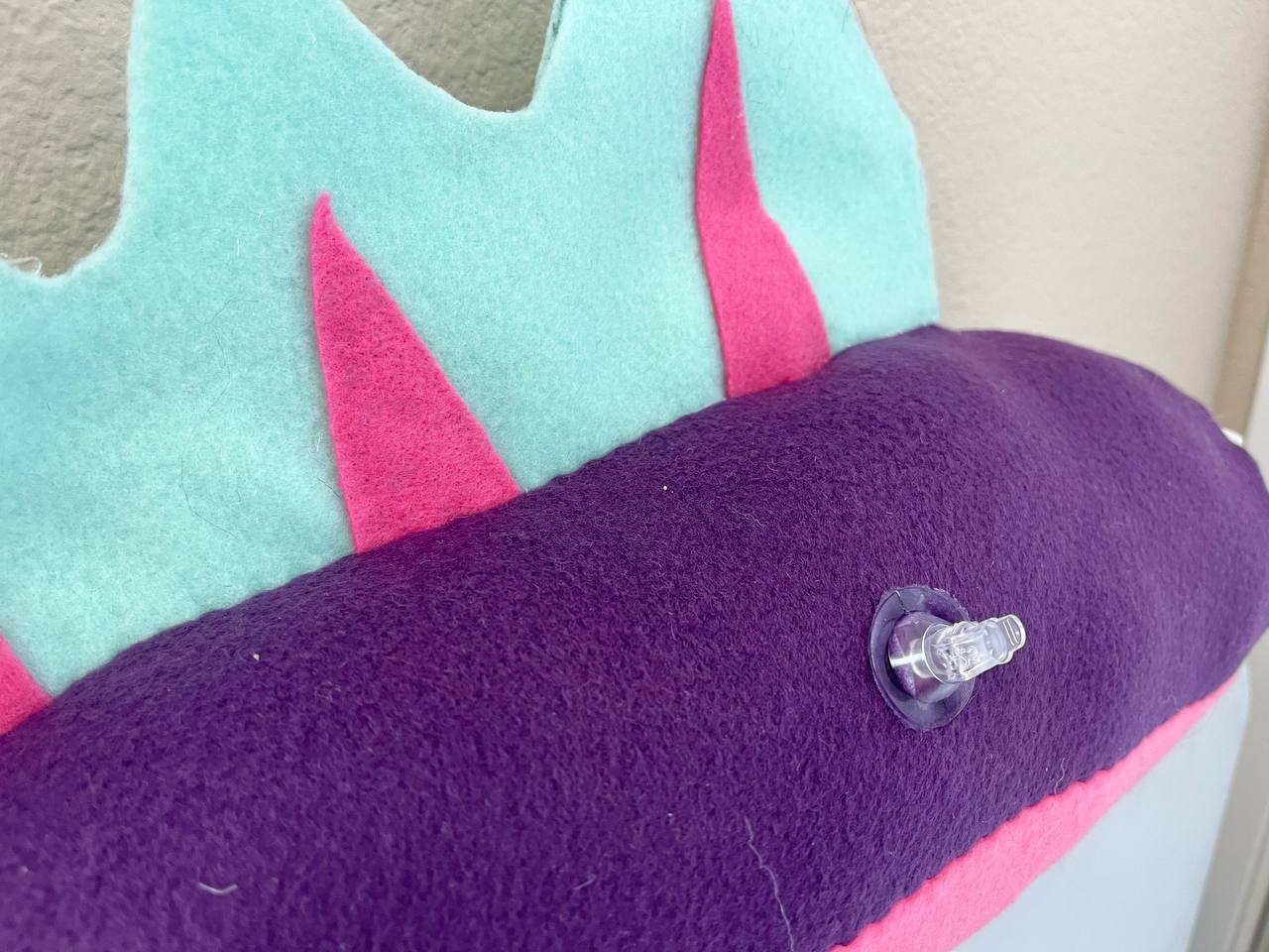 Picture of a Pooltoy inspired fursuit tail, made by Radio Neyo. It is mainly purple with a pink underside and light blue and pink fins on the top. 